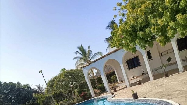 A swimming pool with a tiled edge, surrounded by a paved area and a white building with arches.