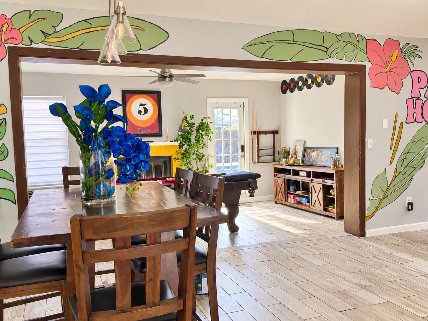A dining area with a wooden table and chairs, a vase with blue flowers, and a wall with a tropical mural.
