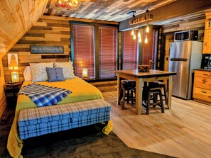 A cozy bedroom with a bed, bedside table, and a nightstand. There is a kitchen area with a refrigerator, microwave, and bar stools.