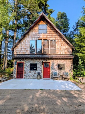 A two-story cabin with a stone facade, a red door, and a small porch with outdoor seating.