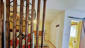 A room with a bed, a wooden floor, a wall with a clock, and a door leading to another room. - GOZO APARTMENT (Qala)