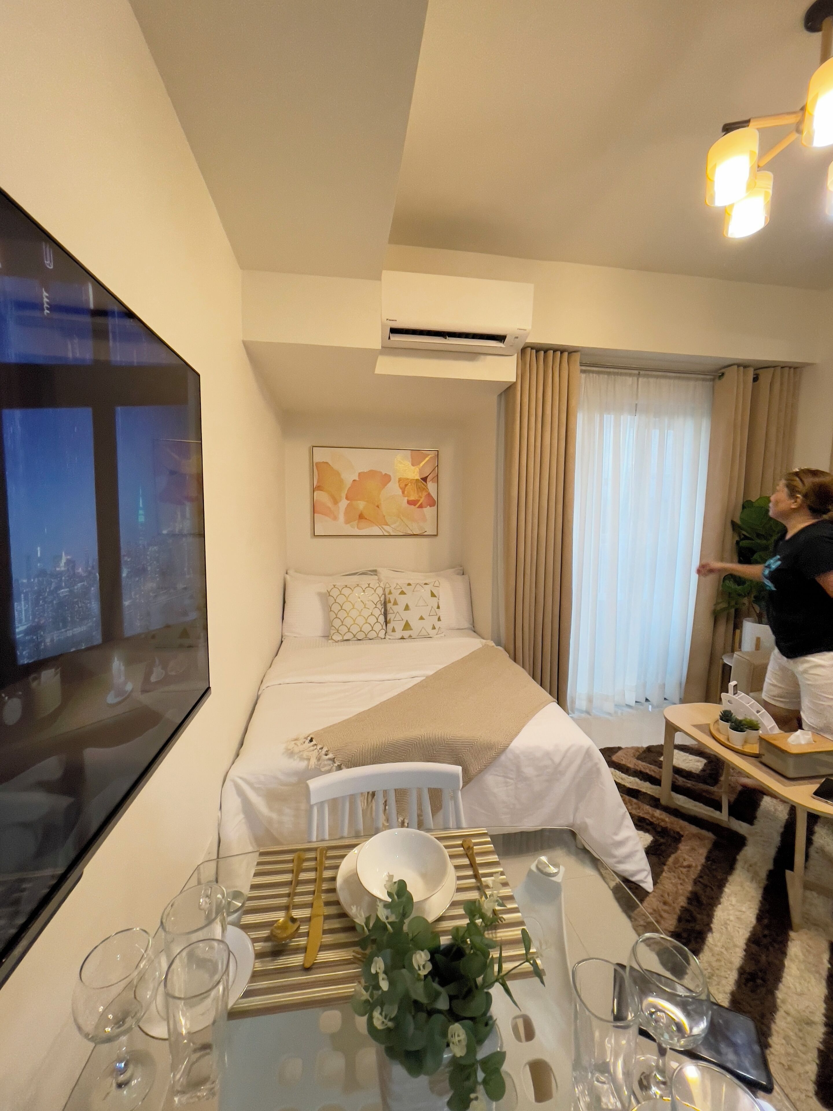 Ein Hotelzimmer mit einem Bett, einem Fernseher, einem Tisch mit Gläsern und einer Pflanze sowie einem Fenster mit Vorhängen.