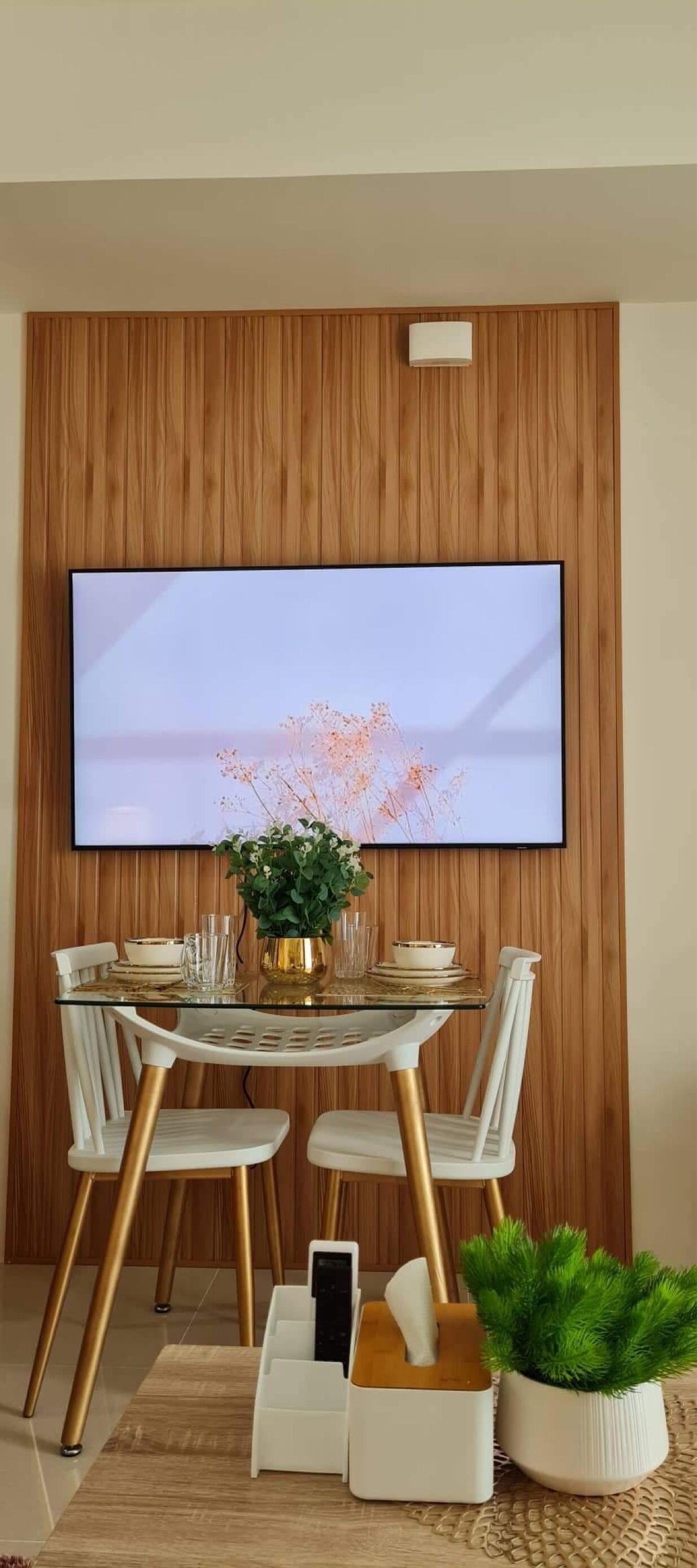 Un comedor con pared de madera, televisor de pantalla plana, mesa de vidrio, sillas blancas y una pequeña planta en maceta.