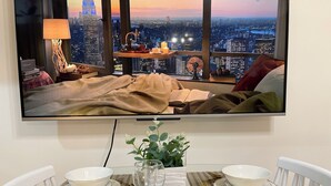 Quarto com vista para a cidade, uma cama com cobertor, mesa de cabeceira com luminária e televisão na parede.