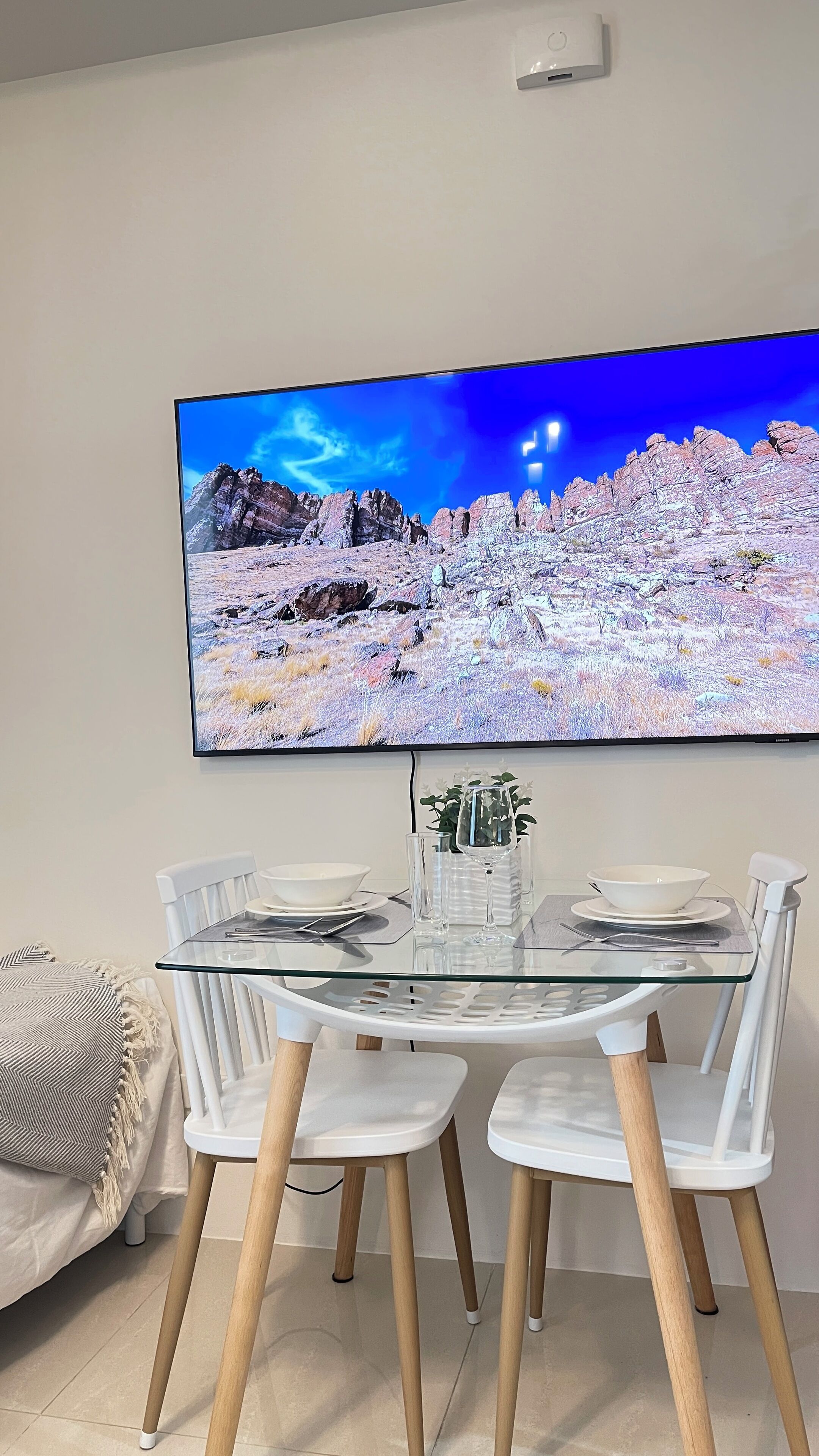 A dining area with a glass table, white chairs, and a flat-screen TV displaying a mountainous landscape.