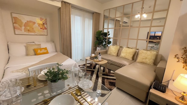 Quarto de hotel moderno com cama, sofá, mesa de jantar posta para dois e uma janela grande com cortinas.