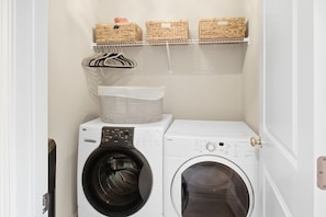 A laundry room with a front-loading washing machine and dryer, a shelf with baskets, and a hanging rack. - New 3BR Home - Walk to Downtown Belmont - Near CLT - Sleeps 8 (Belmont)