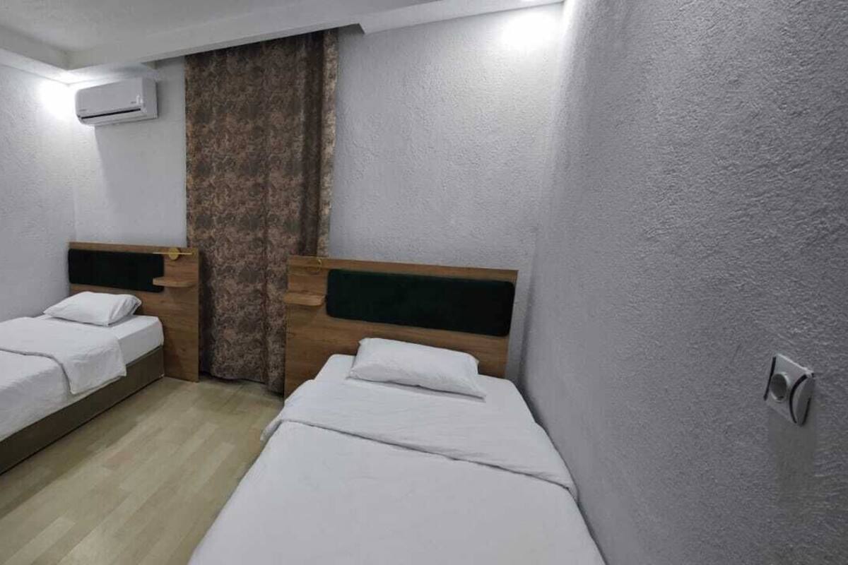 Una habitación pequeña y limpia con dos camas individuales, un cabecero de madera, aire acondicionado de pared y una cortina.