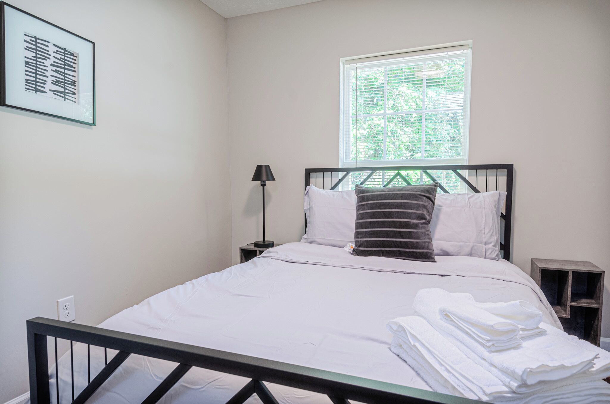 A neatly made bed with white linens and a black metal frame, a bedside lamp, and a window with blinds.