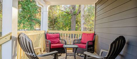 A covered outdoor seating area with wicker chairs and a red sofa, surrounded by a wooden deck and trees.