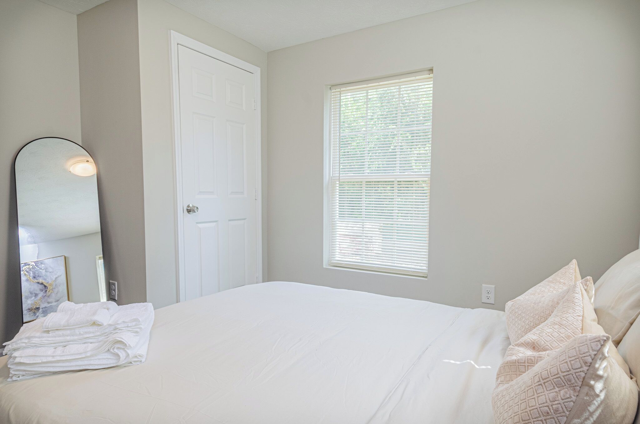 A neatly made bed with white linens, a white door, and a window with blinds.