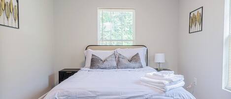 A neatly made bed with a headboard, a bedside table with a lamp, and a window with blinds.