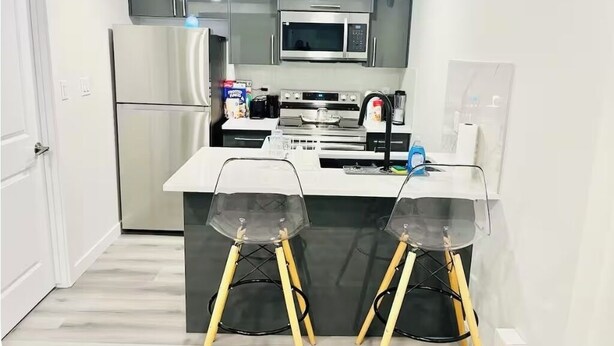 A modern kitchen with a refrigerator, microwave, stove, and two clear plastic chairs with wooden legs. - Stylish One Bedroom Cozy Home (Edmonton)