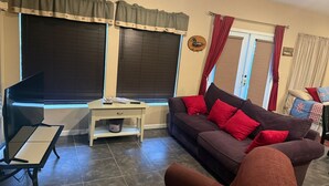 A living room with a dark brown sofa, red cushions, a white side table, a television, and a window with blinds.