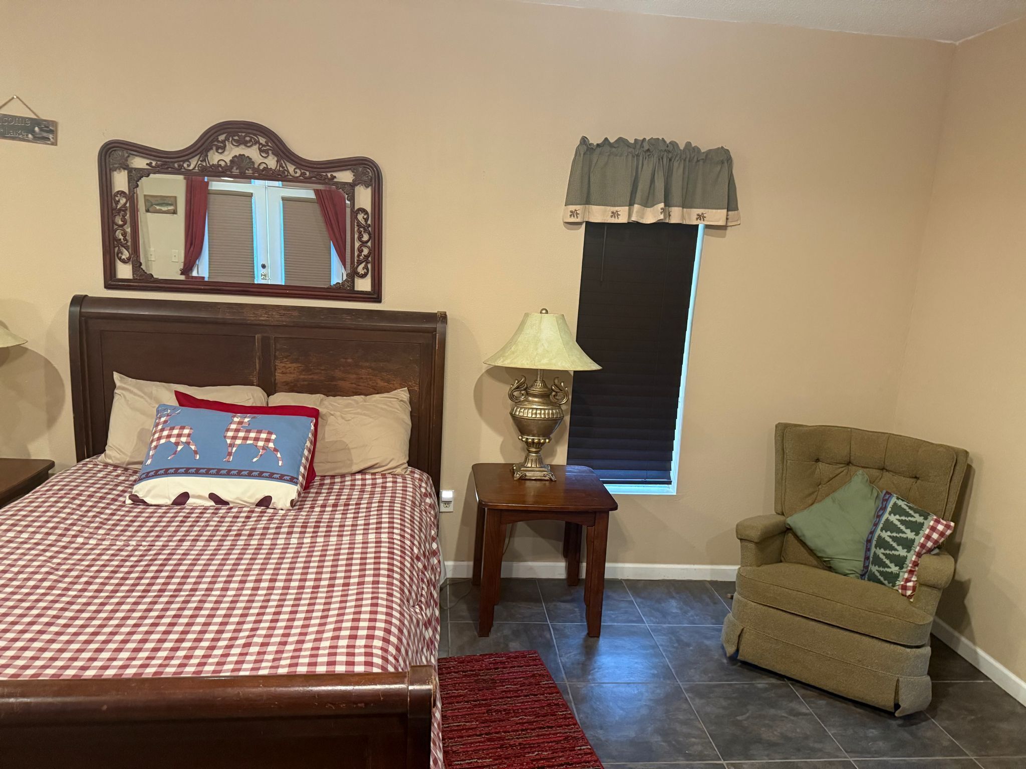 Ein Schlafzimmer mit einem Bett, einem Nachttisch, einer Lampe, einem Sessel, einem Fenster mit Jalousien und einem Spiegel an der Wand.