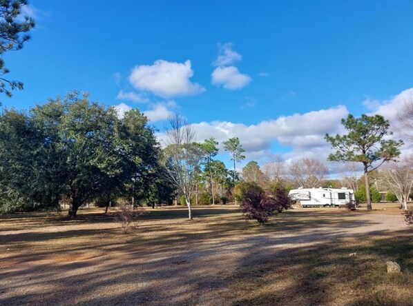 A campground with a white RV in the distance, surrounded by trees and open grassy areas.