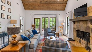 A cozy living room with a fireplace, wooden ceiling, and large windows. - Firefly Cottage, Firefly Cottage | Lake Martin Getaway | Sleeps 8 | Pet Friendly (Elmore County)