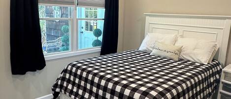 A bedroom with a checkered bedspread, a white headboard, and a window with black curtains.