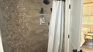 A shower with a pebble wall, a white shower curtain, and a small shelf with toiletries.