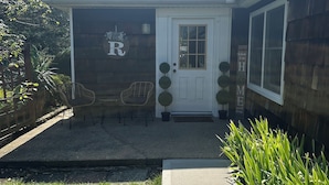 A house with a covered porch, a white door, and a small garden area with chairs and potted plants.