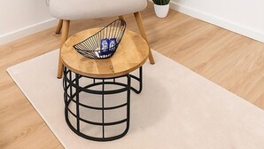 Una habitación minimalista con un sillón de color claro, una pequeña mesa de madera con base de metal, una alfombra blanca y una planta en maceta.