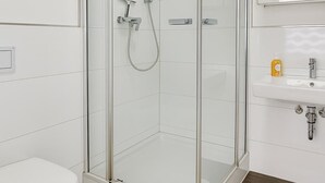 Un baño moderno con ducha envuelta en vidrio, inodoro blanco, lavabo con espejo y dos alfombras de baño color beige.