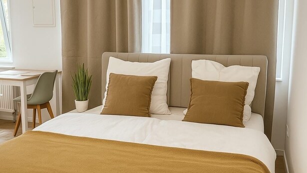 Un dormitorio con cama, cortinas color beige, una planta en maceta y vistas al exterior.