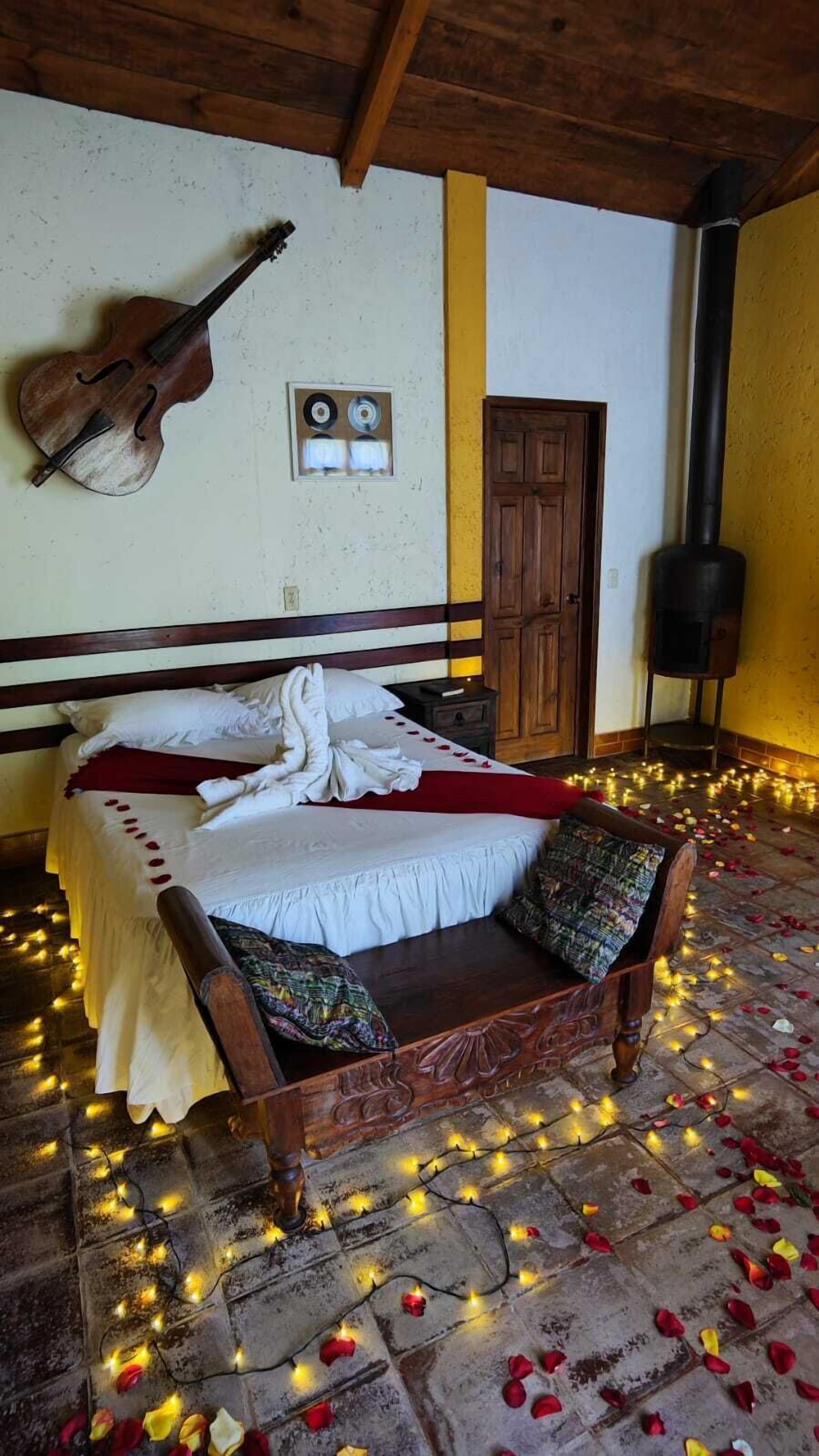 Una habitación con una cama decorada con pétalos de rosa y luces de cadena, una guitarra en la pared y un banco de madera rústica.