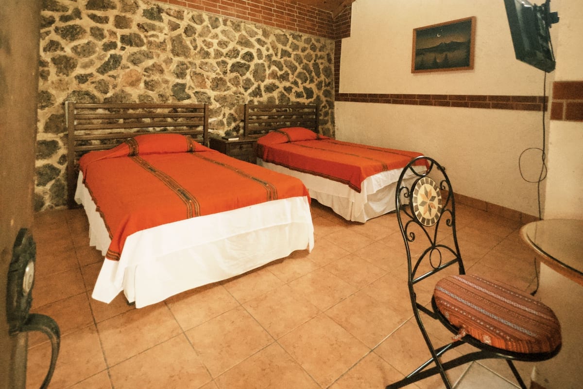 A room with two beds, a stone wall, a wooden ceiling, and a small table with a chair.