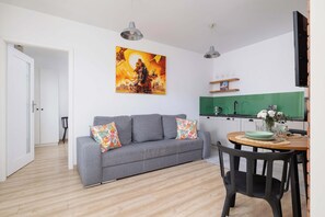 Uma sala de estar moderna com um sofá cinzento, uma mesa de jantar com cadeiras, uma cozinha e um quadro grande na parede.