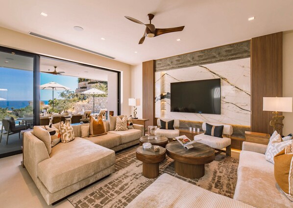 A spacious living room with a large sofa set, a coffee table, and a TV mounted on the wall. There is a ceiling fan and a view of the outdoors through large windows. - New to VRBO, New modern luxury home in Palmilla, pool/spa, swimmable beaches. (San Jose Del Cabo)