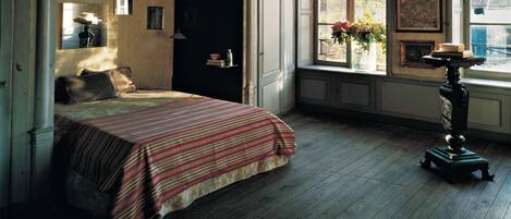 Un dormitorio con cama, una ventana con flores, una lámpara y una estantería con libros y un jarrón.