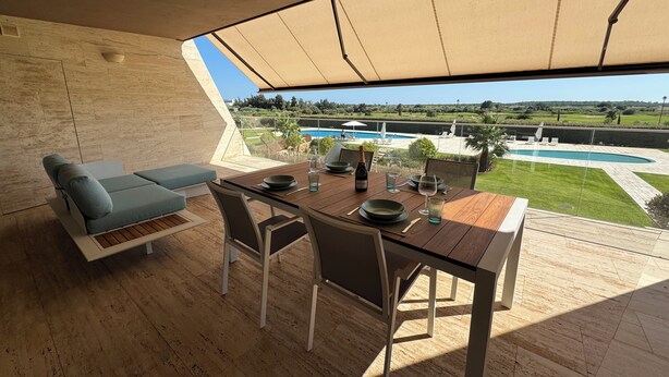 Un área de comedor al aire libre moderna con una mesa de madera, sillas y un banco acolchado. Un amplio toldo proporciona sombra.