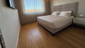 Un dormitorio moderno con una cama grande, una mesita de noche, un televisor montado en la pared y una ventana con cortinas translúcidas.