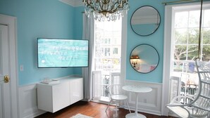 A room with a TV on the wall, a white cabinet, a small round table, a white chair, and a large window with a view of buildings.