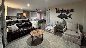 A modern living room with a dark leather sofa, a round wooden coffee table, and two armchairs. A kitchen with white cabinets and a dining area is visible in the background.