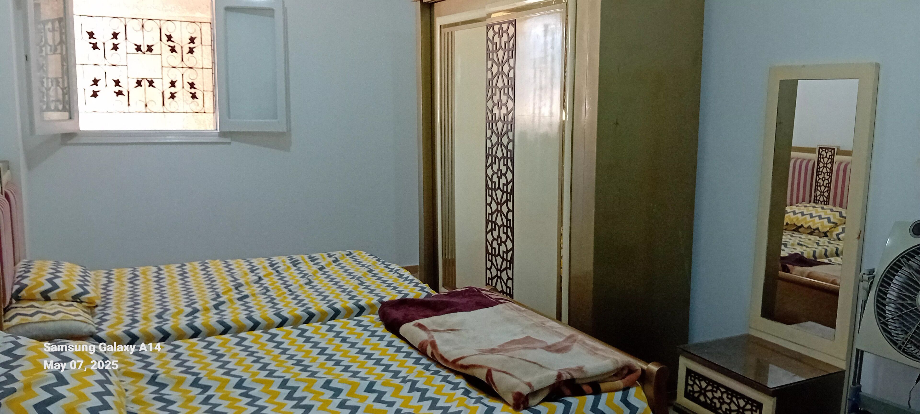 Ein Schlafzimmer mit Bett, Fenster mit Ziergitter, Spiegel und Ventilator.