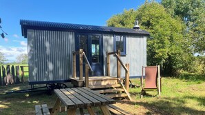 A small, single-story building with a corrugated metal roof and a wooden deck in front. - Cwt Clyd-Shepherd’s Hut (Bangor)