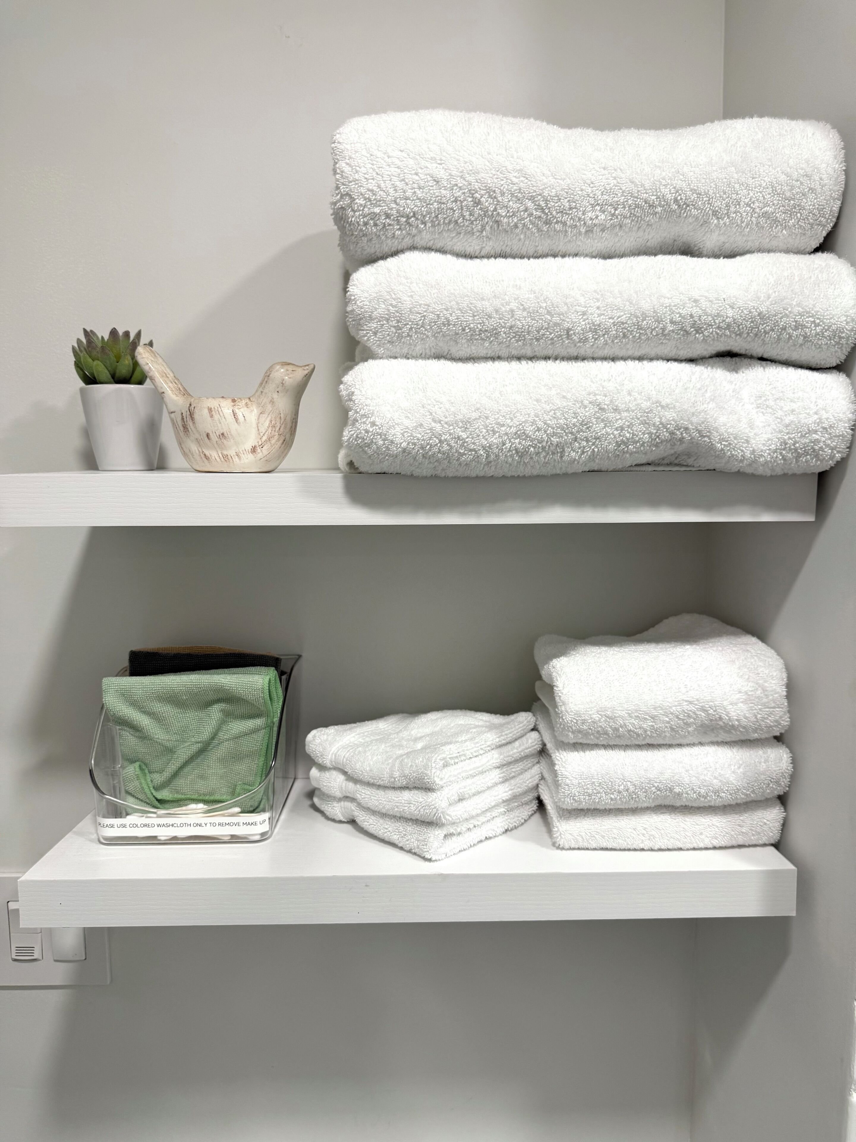 A bathroom with white towels stacked on shelves, a green towel dispenser, and a small potted plant.