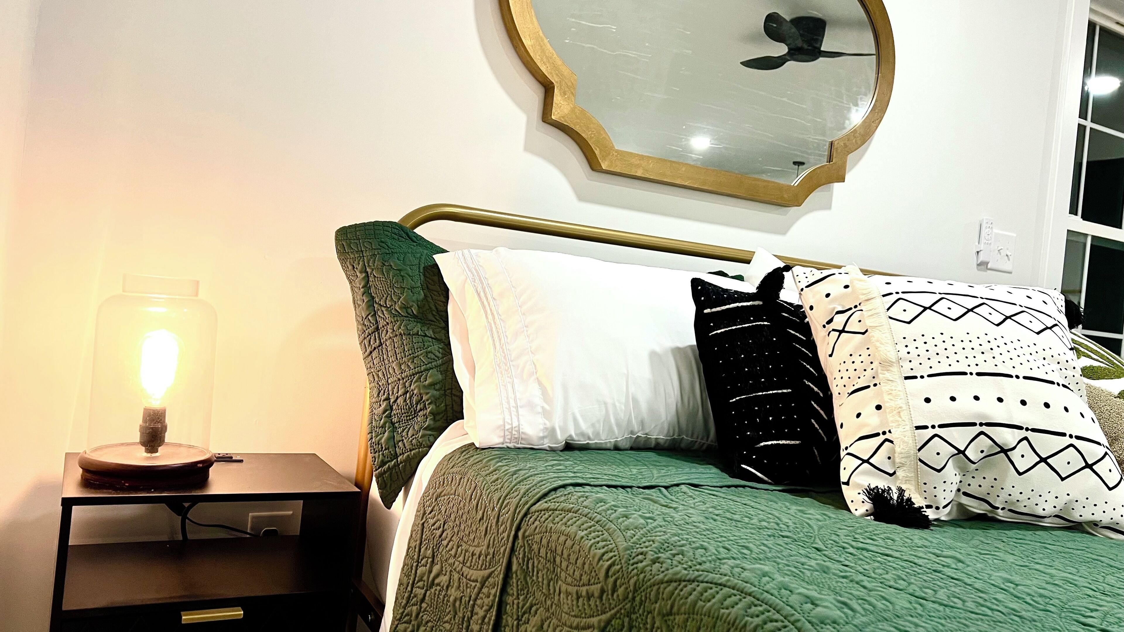 A neatly made bed with green bedding, a bedside lamp, and a mirror on the wall.