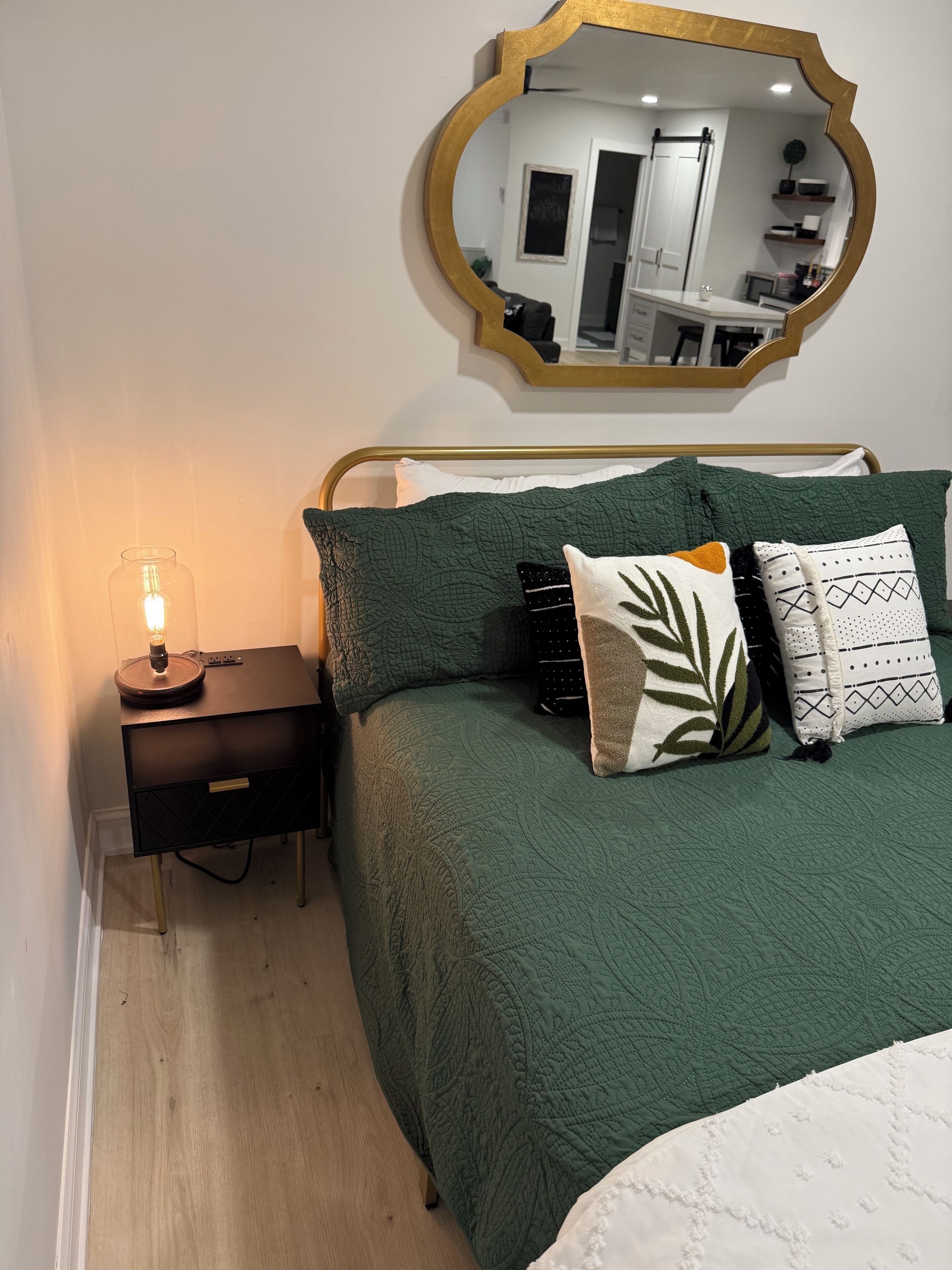 Un dormitorio con colcha verde acolchada, una mesita de noche con lámpara y un espejo en la pared.
