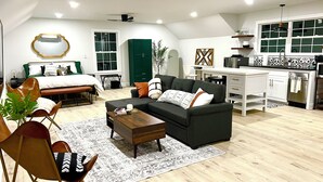Un salon moderne comprenant un canapé, une table basse et des fauteuils, une chambre séparée avec un lit, une cuisine avec des meubles blancs et un plan de travail noir, et un coin repas avec une table et des chaises.