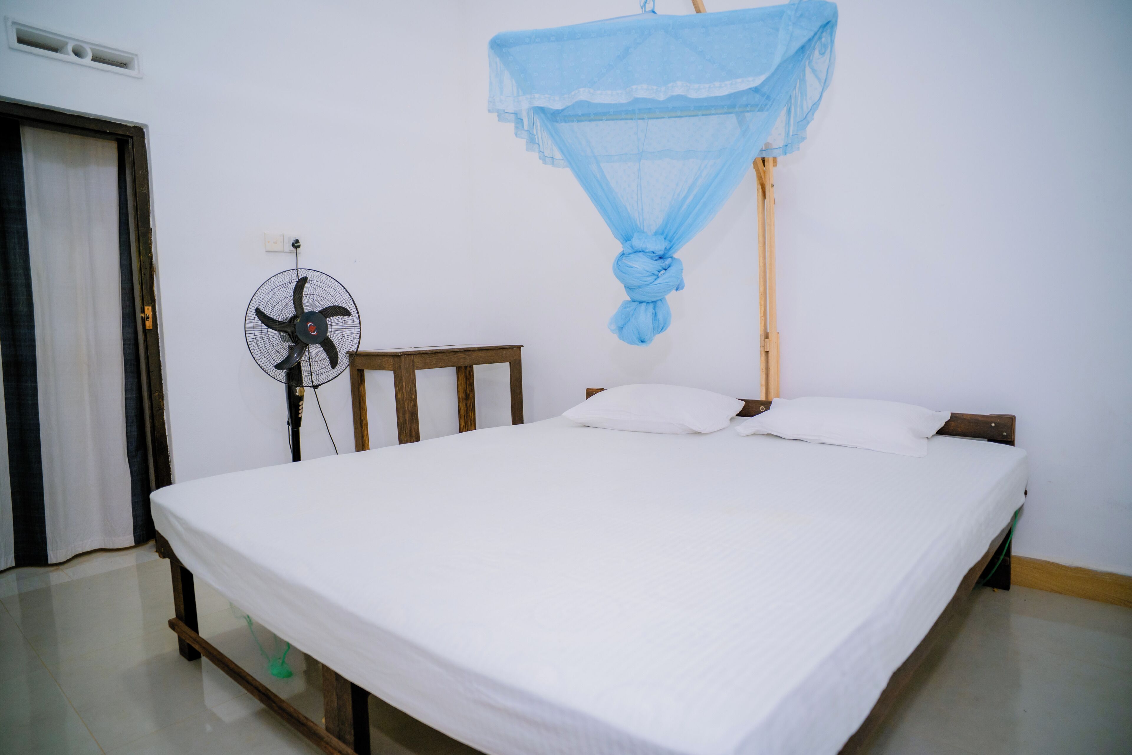 Ein Einzelbett mit weißer Matratze und Kissen, ein hölzernes Kopfteil, ein Standventilator und ein Moskitonetz, das über dem Bett hängt.