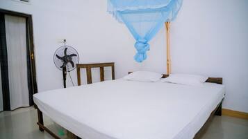 Ein Einzelbett mit weißer Matratze und Kissen, ein hölzernes Kopfteil, ein Standventilator und ein Moskitonetz, das über dem Bett hängt.