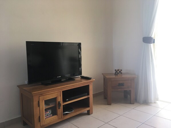 A living room with a wooden entertainment unit holding a flat-screen TV, a remote, and a small cabinet with books. There's a wooden side table with a decorative item on it, and a window with sheer curtains. - Holiday Home Paphos - Near Amenities - Poolside (Paphos)