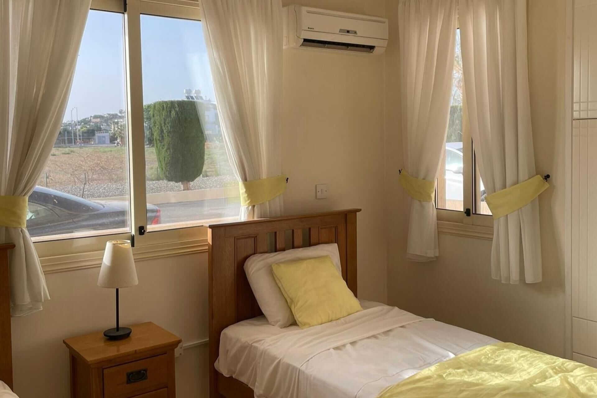 A bedroom with a wooden bed, white bedding, a bedside lamp, and an air conditioning unit.