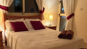 A neatly made bed with white and red bedding, a wooden headboard, and two rolled towels on the foot of the bed. - Holiday Home Paphos - Near Amenities - Poolside (Paphos)