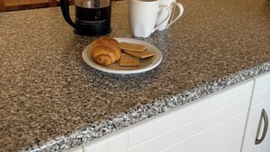 A kitchen counter with a coffee maker, two white mugs, a plate with a croissant and cookies, a wooden chair, and a wall-mounted air conditioner. - Holiday Home Paphos - Near Amenities - Poolside (Paphos)