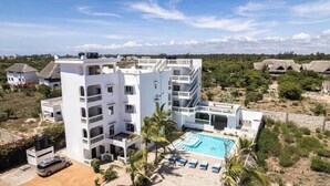 Um hotel branco de vários andares com piscina, rodeado por vegetação e outros edifícios.