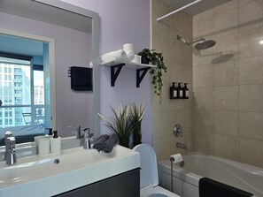 Une salle de bain moderne avec un grand miroir, un lavabo avec des articles de toilette, des toilettes, une douche avec un pommeau de douche à main et une fenêtre donnant sur des immeubles.